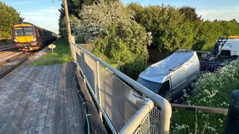 Cambridgeshire Police View from the top of the railway bridge shows the van being recovered from under the bridge. The roof is dented and crumpled inwards. The van is being towed away from behind. A train is stopped on top of the railway bridge while recovering is underway.