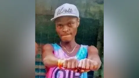 Jecinta Gathoni A young man with a baseball cap and a tie-dyed vest holding his fists in front of him.
