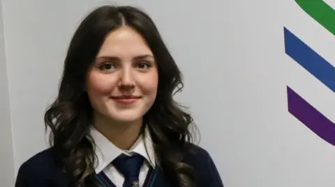A girl with dark hair is standing in front of a white wall with her school logo to her right. She is wearing a navy blue school uniform.