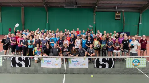 Swindon Pickleball A crowd of people posing for a photograph in an indoor pickleball court. A net is in the foreground and a ball is on the floor.