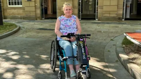 Flick Williams Flick Williams sat in a wheelchair, wearing jeans and a pink flowery top, with short blonde hair. She is pictured outside a building.