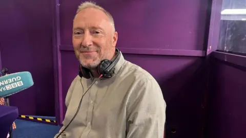 A man with short grey hair smiling at the camera. He has over ear headphones around his neck. You can see a microphone branded with BBC Radio Guernsey to the left.