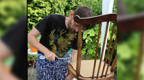 Huntington's Disease Association A bearded man hammering nails into a dining room chair to secure a cushion.