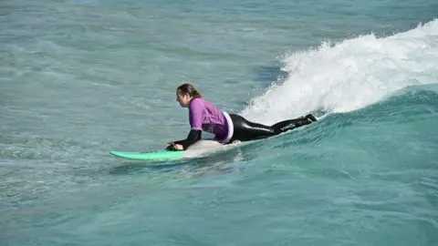 Kay Millar Kay Millar surfing a wave on her green surf board. She is pictured in the prone position on the board and is wearing a purple rash vest over a black wet suit.