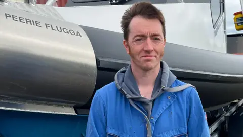 Close-up of a person wearing a blue work jacket standing beside a large rigid inflatable boat on a trailer, with the name “PEERIE FLUGGA” visible on the vessel and industrial buildings in the background.