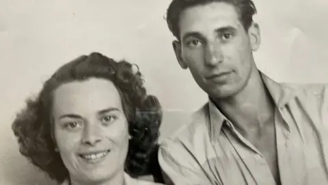Bolton Family Black and white picture of a couple with a white background the lady on the left has dark black hair and a light coloured top on the man on the right also has dark hair swept across his head and an open topped light shirt.