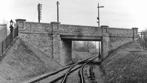 Buntingford Railway & Local History Society collection A black-and-white photograph shows a railway line curving gently as it passes beneath a substantial brick bridge. Steep grass-covered embankments rise on both sides of the track, creating a cutting through the landscape. Traditional telegraph poles and railway signalling equipment are visible above and alongside the bridge.
