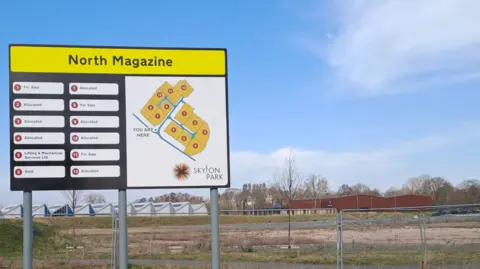 A sign denotes the firms on the enterprise zone. It stands in front of wasteland ready for development. The words North Magazine are in black font on a yellow background at the top of the sign.