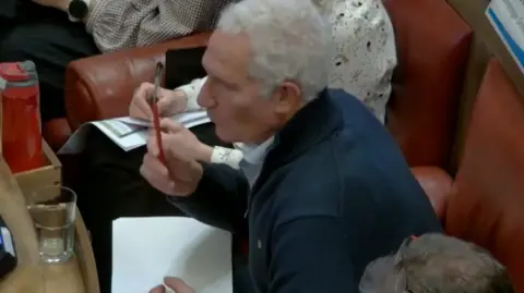 Cambridge City Council An image from a council meeting livestream of Peter Freeman, who is talking. He has short white hair and is wearing a navy jumper and sitting on a red leather chair. The image is taken from above.