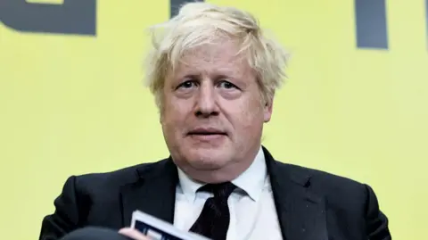 Getty Images Boris Johnson has unkempt blonde hair and is wearing a black jacket, black tie and white shirt. He is sitting in front of a yellow backdrop