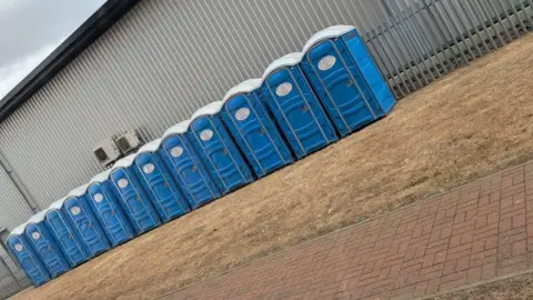 Supplied A row of blue portable toilets, by a metal building, with an area of grass in front and a brick path. A metal railing is to the right. 