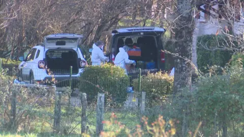 France Télévision Forensics officers taking boxes either to or from the boots of a car, with another car with its boot open next to them. Trees and bushes are all around them.