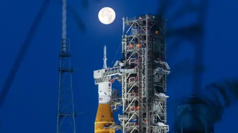 A large and very bright full Moon appears directly on the top of Nasa's Space Launch System rocket at night. The orange and white rocket is sitting next to a steel support structure called a mobile launcher.
