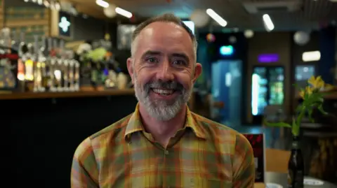 Hannah Karpel/BBC Man in the foreground wearing a yellow and green checked shirt, seated indoors and facing the camera. The setting is a bar or pub, with a row of beer taps and bottles visible behind the person on the left. Tables and chairs are positioned in the background, and a small vase containing yellow flowers sits on a table to the right. Overhead ceiling lights and hanging decorations are visible throughout the venue. No bags, bins or markers are visible.