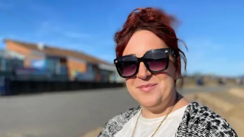 Cara Swarbrick stands on a promenade on the Lincolnshire coast. She has dark-red hair, tied back, and is wearing big sunglasses, a white T-shirt, a gold necklace and a black and white cardigan.