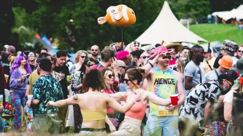 Goodgroove A crowd of people wearing colourful summery clothes at a festival. Two women have their arms stretched out and are dancing. There is an orange clownfish toy being held up on a stick by someone in the crowd. A small domed tent is in the distance.