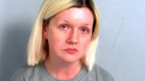 Essex Police Custody mugshot of Virginia McCullough. She has light blond hair with dark roots. She is wearing a grey sweater and is looking solemnly at the camera.