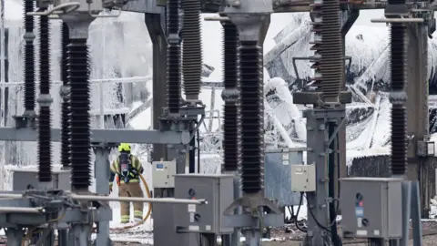 Getty Images A uniformed firefighter sprays foam onto parts of the substation on Friday morning. White foam covers many parts of the substation in the background