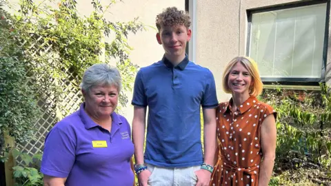 Royal United Hospitals Bath NHS Foundation Trust A teenager standing in the middle of the photo, next to his mum and a hospital worker. The three are photographed outside with plants behind them