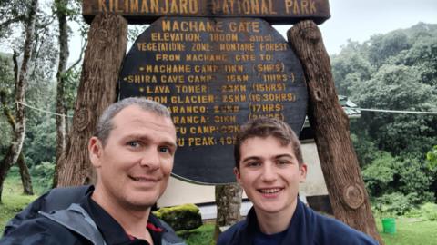 A selfie of a man with grey hair, black shirt and navy coat and a young boy with brown hair and a navy jacket. They are both standing in front of a brown wooden sign saying Kilimanjaro National Park