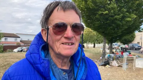 A man with dark, graying hair, wearing sunglasses and a blue bomber jacket stands in the foreground. A green space with trees is behind him. Around the trees is several different piles of waste.