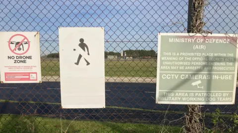 Matt Precey/BBC Three large signs sit on a fence warning people that the area is a no drone zone and there is a prohibited space behind the fence. An airbase sits behind the fence and several small buildings are seen in the distance. 