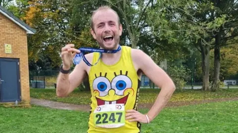 Alex Hunt with short brown hair and beard, He is wearing a medal on a dark blue ribbon around his neck. He is holding the medallion in his right hand, while his left is resting on his hip. He is wearing a yellow running vest, which has a face on the front with large eyes, a small nose and a smiling mouth with two teeth. His number, 2247, is on the vest in black writing. He is standing on grass with trees behind him.
