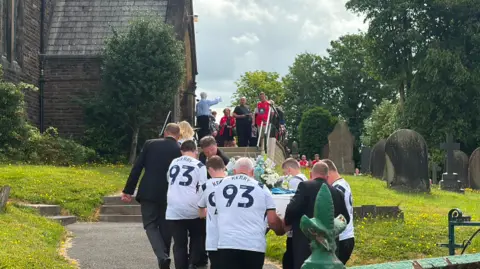 Kerry Coombes's white coffin with white and pale blue flowers on top is carried by mourners dressed in football tops with the number '93' and 'Kerry' up the steps leading to All Saints Church in Higher Walton. It is a sunny day.