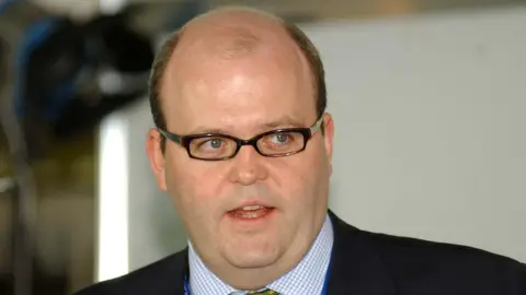 Getty Images A man is speaking. He wears glasses and has a black suit on with a checked blue and white shirt. His tie is green and patterned.