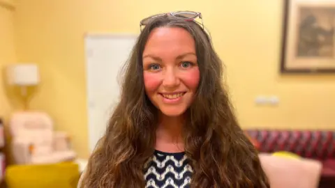 Rebekah Hester smiling at the camera. She has long brown hair and is stood in a lounge with a red sofa in the background and yellow walls.