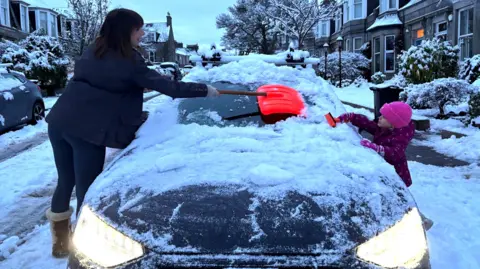 A mother and daughter clear the snow-laden windscreen of a car in the early hours. The car is on an upmarket residential street with sandstone properties, trees and bushes.