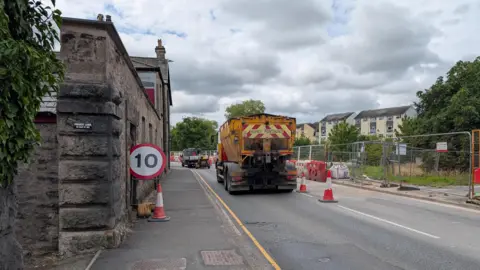 A large lorry driving down Aynam Road in Kendal. One lane is closed and there is a 10mph speed limit sign, white and round with a red edge and digits in black. There are red and white road cones and fencing along the road. There sky is very cloudy.