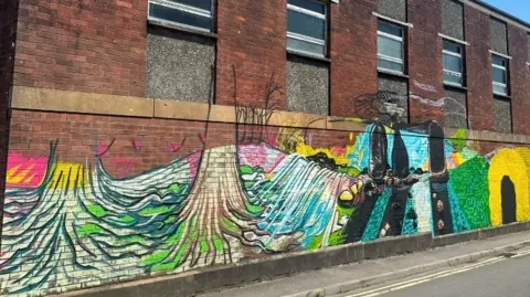 Unfinished paintwork of colorful trees and figures at the bottom of a large brown brick building.