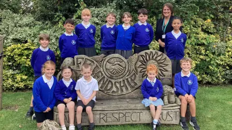 St John's Primary A group of children at St John's Primary in Keynsham sit around a wooden ornament with slogans on it. They are wearing dark blue uniform jumpers and blazers and their teacher is standing with them wearing a black jacket