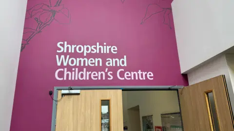 The foyer at the maternity unit at the Princess Royal Hospital in Telford. A sign, on a magenta background, says "Shropshire Women and Children's Centre" and is over wooden double doors, one of which is open.