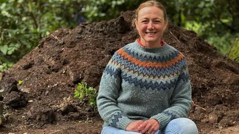 Dr Tori Herridge -a woman with auburn hair and a stripy jumper, sitting at an archaeological dig in a Glasgow park