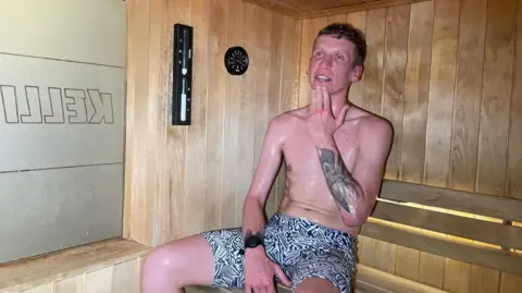 Shaun Whitmore/BBC Logan sitting in a sauna at Kelling Heath in Norfolk, sweating heavily wearing blue and black striped shorts