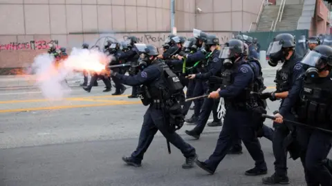 A line of riot police fire weapons as they advance down a street.