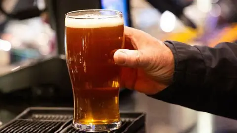 A hand holds a pint of beer in front of pumps at a bar.