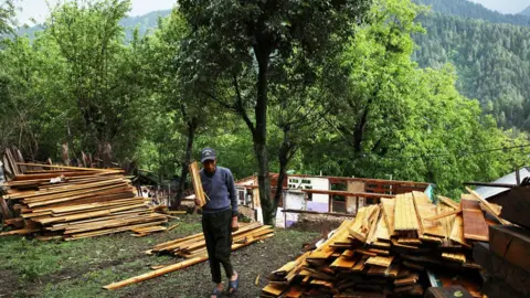 TASEER BEYG / BBC A man in a cap and blue jumper carries a plank of wood, others laid out either side of him and a destroyed house behind him. There is a lush valley in the background.