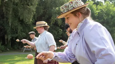 Blists Hill Men and women dressed in Victorian costume take part in a spud and spoon race in a Victorian sports event at Blists Hill. The women are wearing long skirts, which they are holding up with one hand, and blouses rolled up at the sleeves, with straw hats and ribbons. The men are wearing trousers and shirts and flat caps. It is a sunny day and the event is on grass with trees behind them.