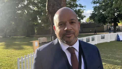 John Devine/BBC Quinton Green is wearing a navy suit and a dark red tie - he has a close cut beard