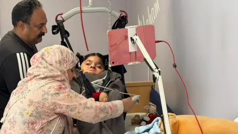 Image shows Nomia's parents operating a hoist to lift her. Her dad wears a tracksuit while her mum wears a floral dress and headscarf