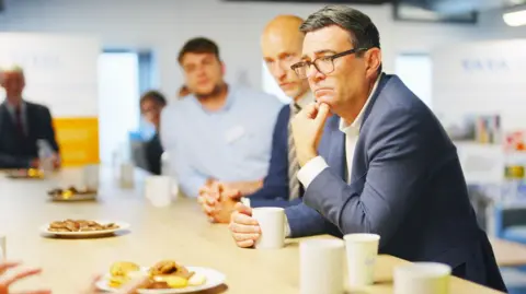 Andy Burnham with Stephen Kinnock sat at a table with bicsuits and mugs. They both have blue suits on. They are in focus, while other people are also sat there who are blurred.