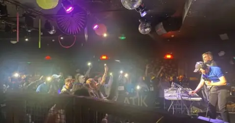 Nue Edmaster and Frosty were among the competitors at the Atik in 2023. The two men are on stage. The man in front is working on computer equipment and has headphones on and his holding a microphone. The performers are watched by crowd of people, some holding their phones up to record so the lights of the phones can be seen.