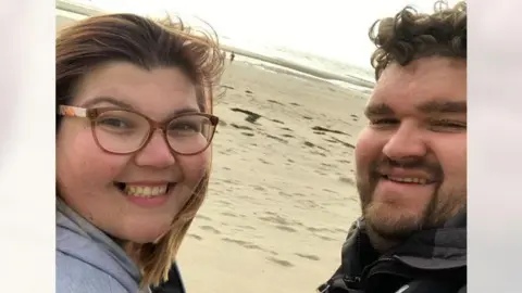 PA Media Gabby Evans and Tom Brakewell are smiling in a selfie taken on a beach. Gabby is wearing glasses.