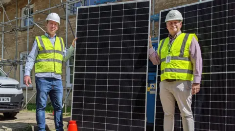 Cotwold District Council Simon Godfrey, Managing Director of Ecovision and councillor Mike McKeown, standing either side of a large solar panel and holding the sides