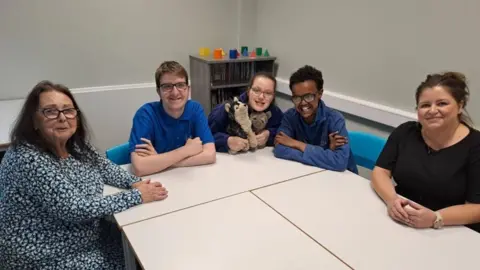 A woman with patterned dress, shoulder length brown hair and glasses sitting at a table with three school pupils wearing blue tops and another woman with brown hair tied back and a black top. They are in a classroom setting.