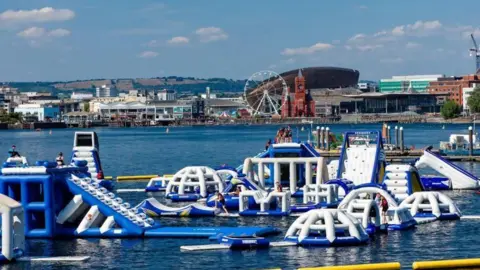 A blue and white inflatable assault course on water with the iconic Cardiff Bay skyline, including a big wheel, the Wales Millennium Centre  and the Pierhead building, in the background.