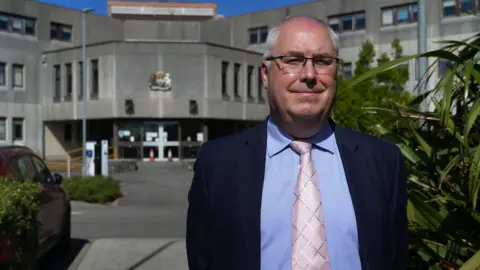 A man wearing glasses stands outside a grey building - the council headquarters. He has white hair and wears a pink tie and a blue suit.